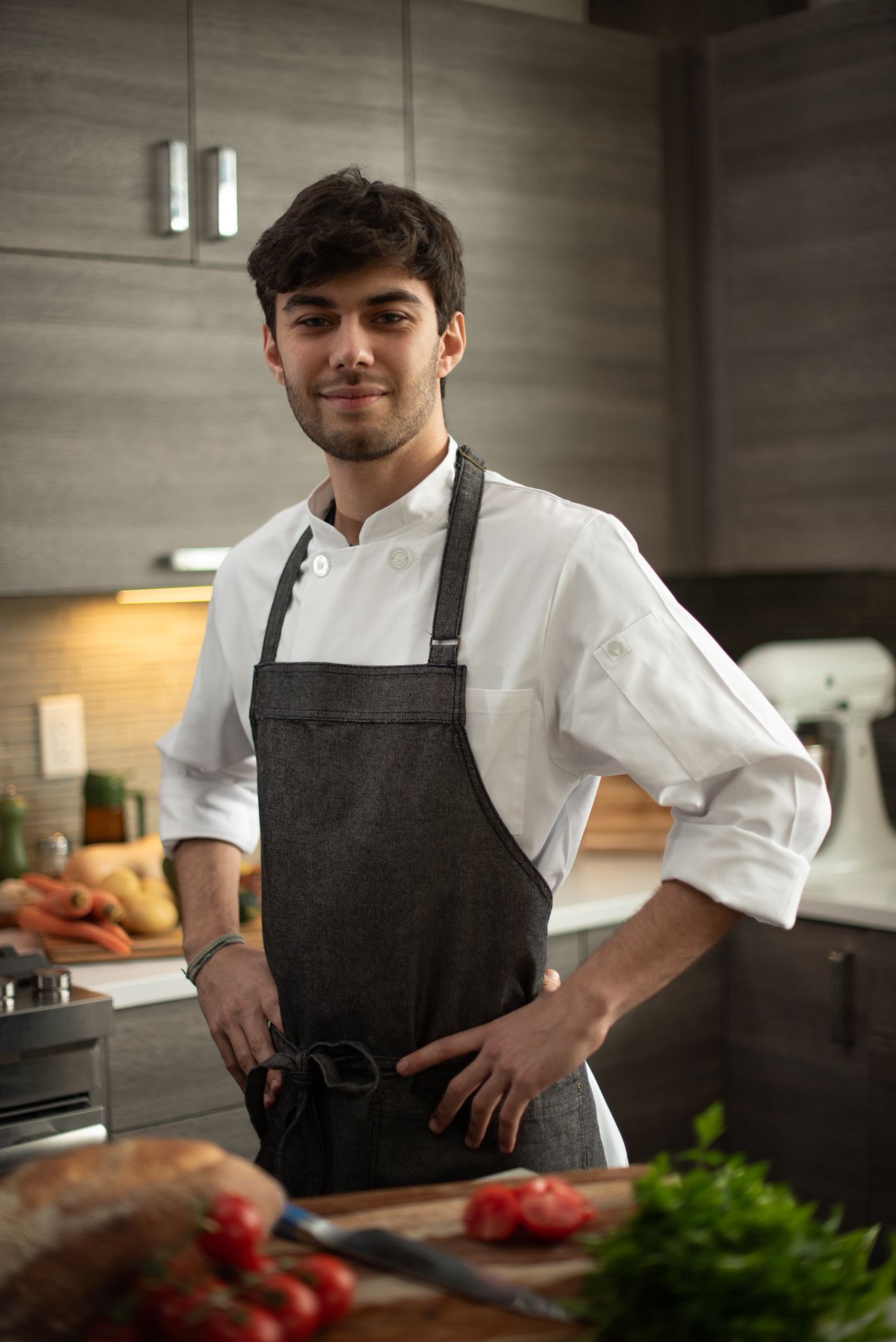 Chef Nicholas Olson in a white chef's jacket and grey apron, hands on his hips, smiling in a kitchen