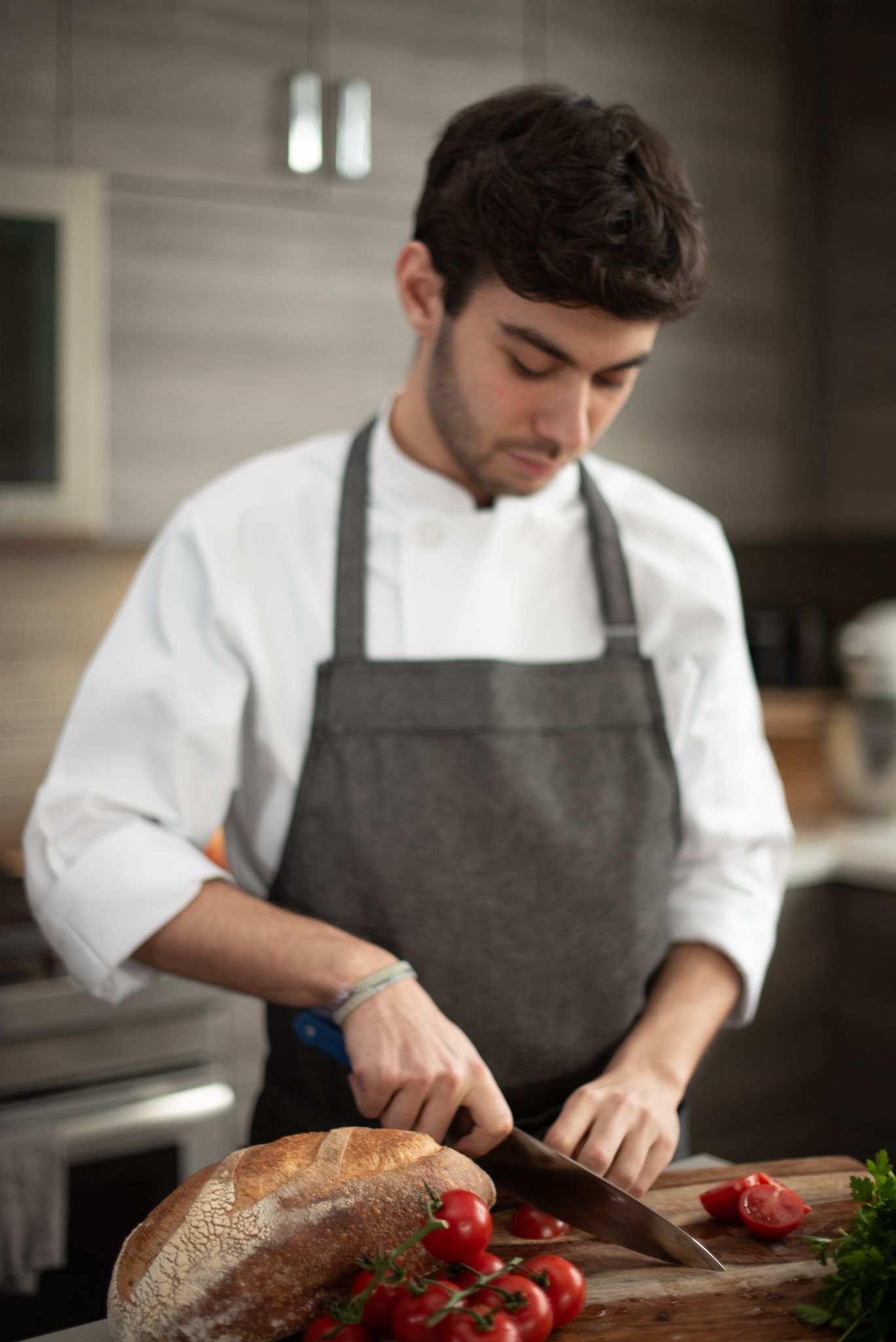 Chef Nicholas Olson in a white chef's jacket and grey apron, slicing cherry tomatoes next to a loaf of bread