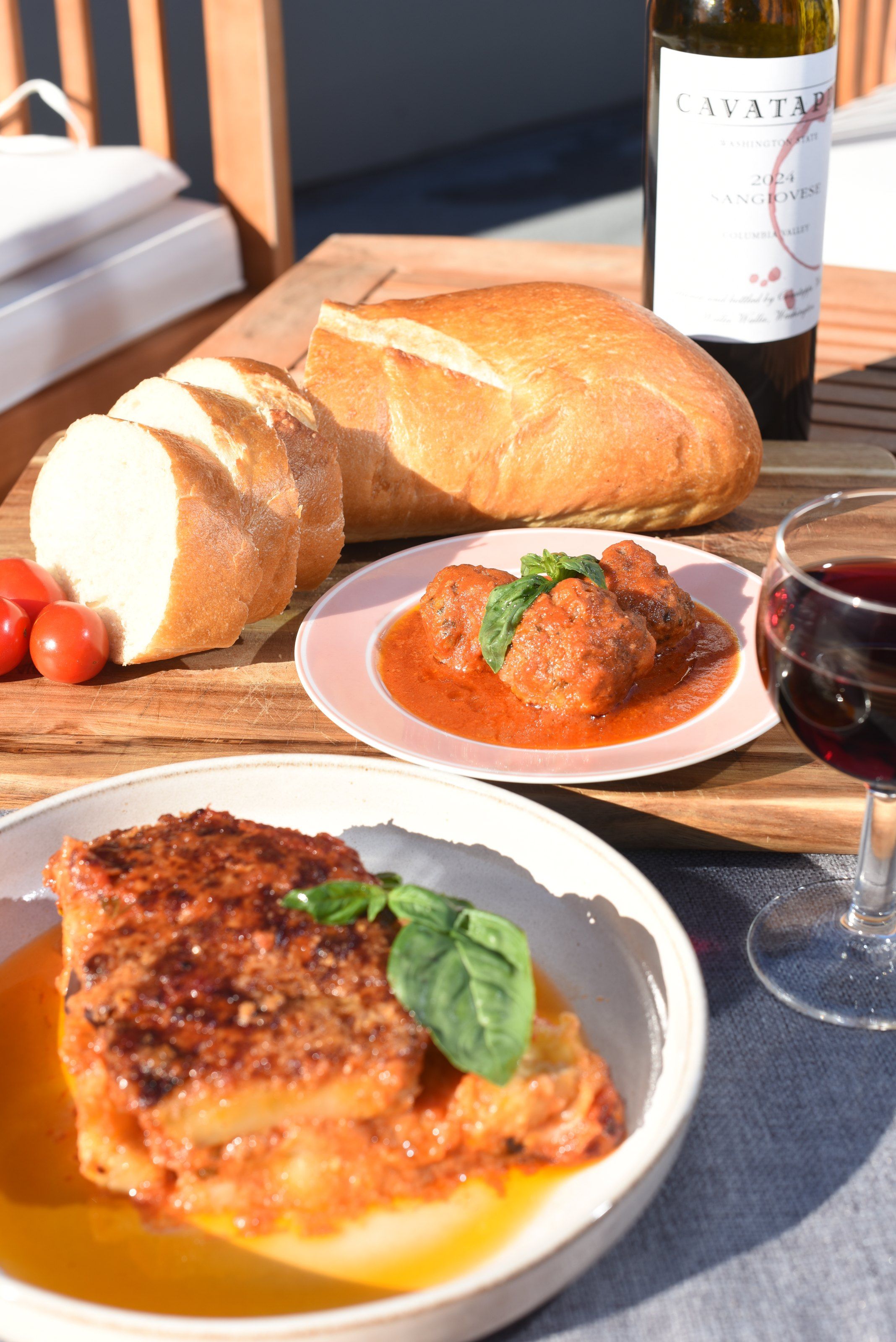 A table laid for two with a square of eggplant parmigiana, a plate of meatballs in tomato sauce with basil, sliced bread, cherry tomatoes and red wine