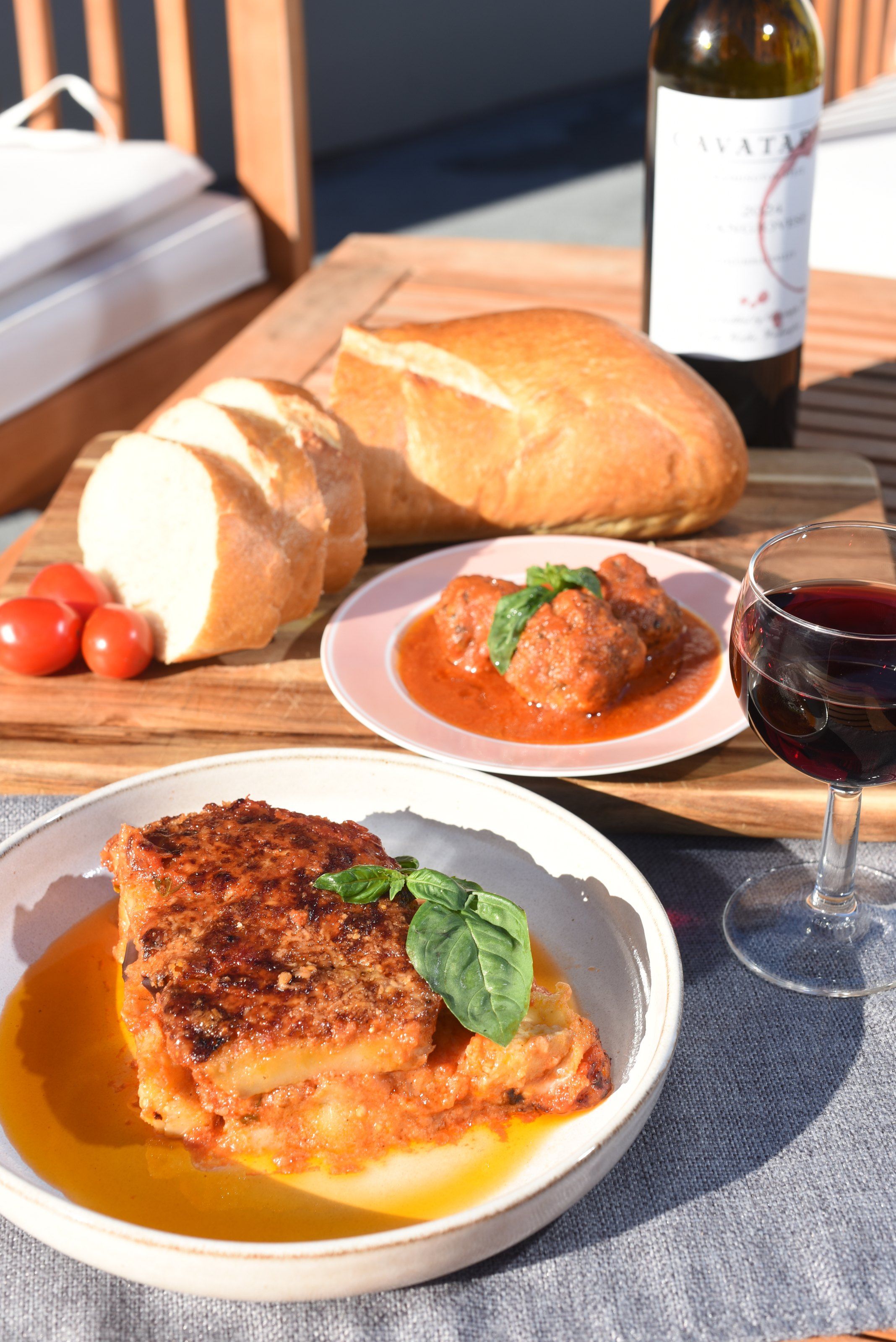 A table laid for dinner with eggplant parmigiana, a plate of meatballs in tomato sauce, sliced bread, cherry tomatoes and a bottle of red wine