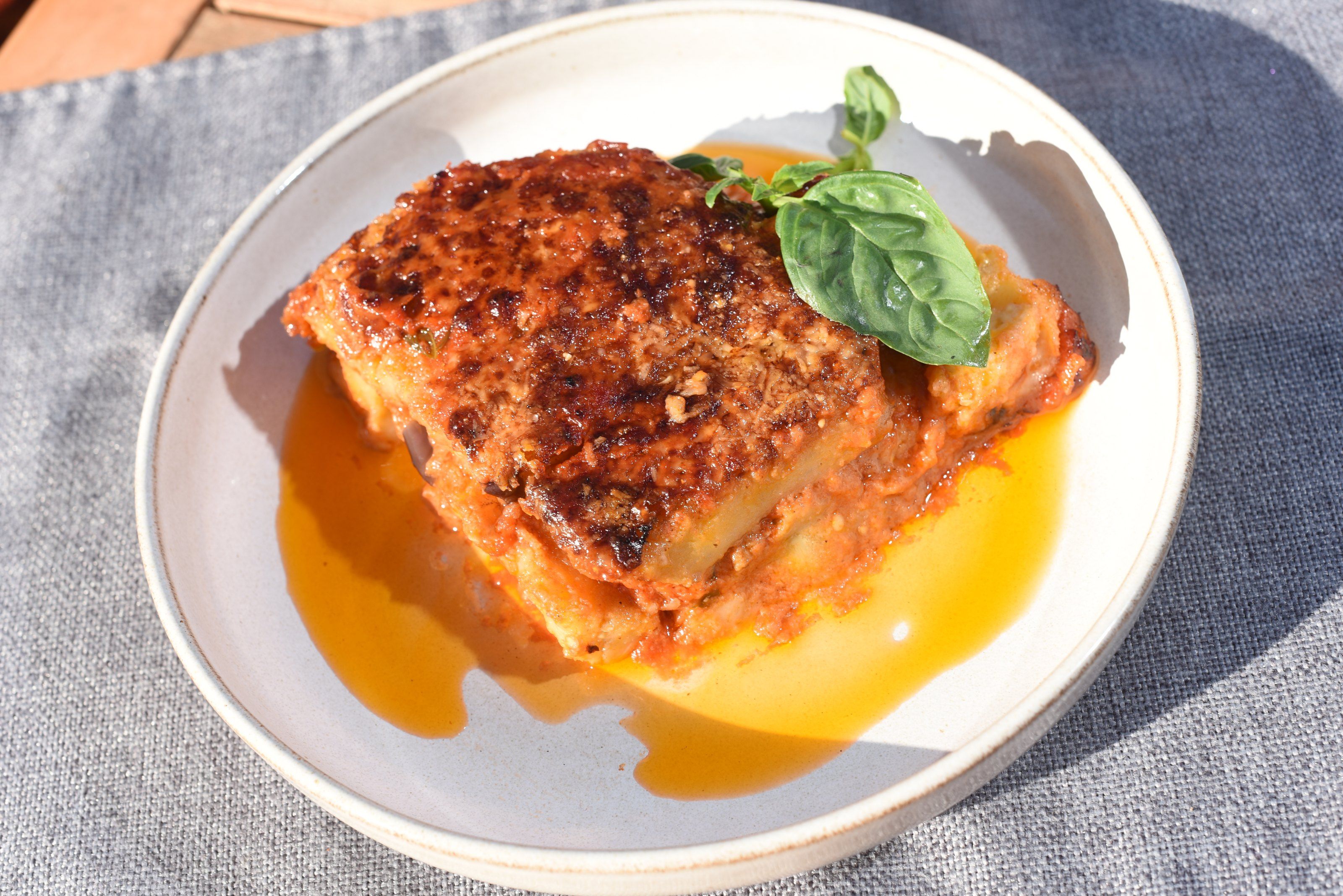 A square of eggplant parmigiana with a basil leaf, in a shallow white bowl on a grey linen placemat
