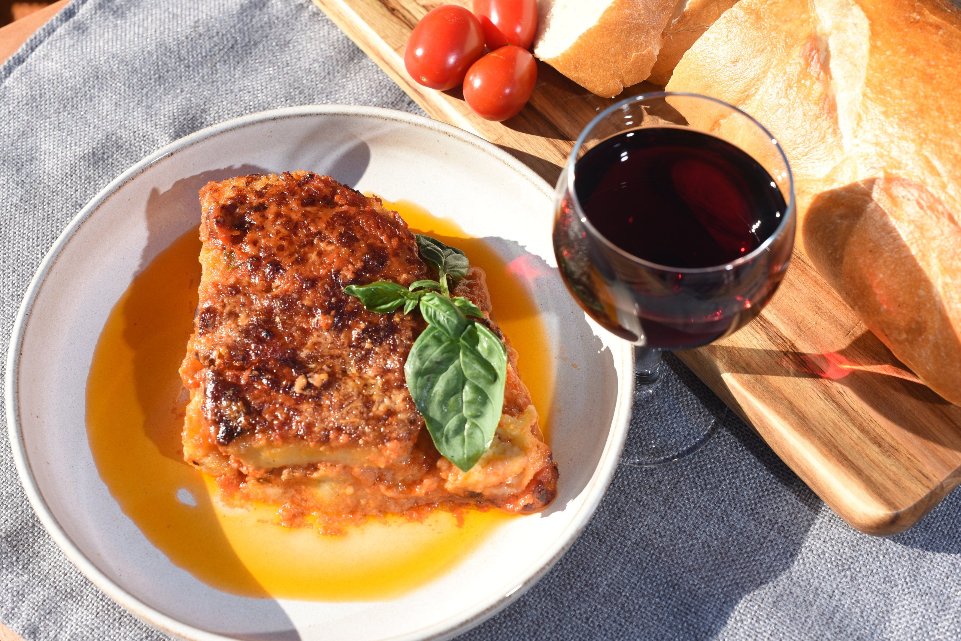 A portion of eggplant parmigiana with basil, a glass of red wine and sliced bread, seen from above