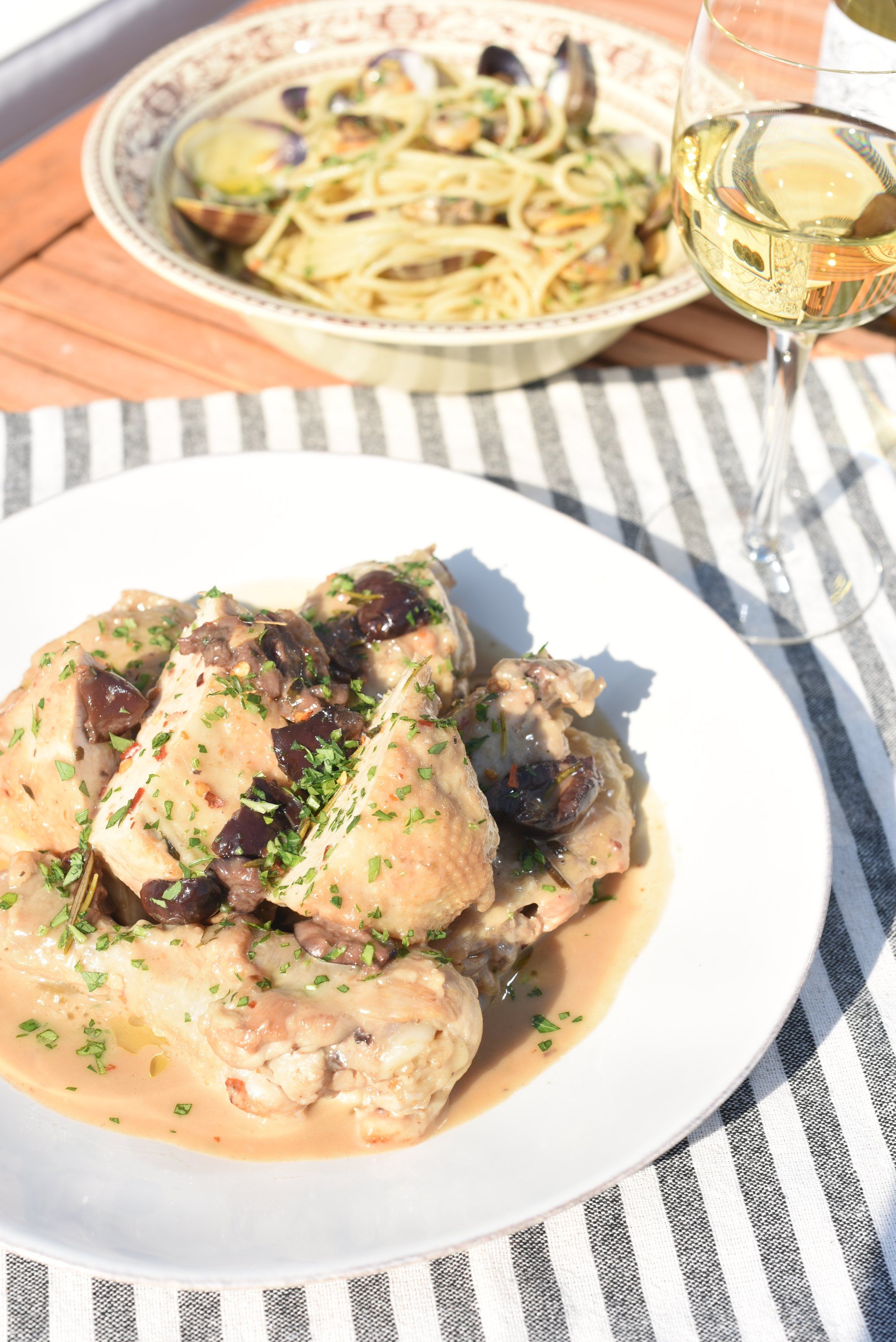 Chicken cacciatora with olives and parsley on a white plate, with a bowl of spaghetti with clams and a glass of white wine behind