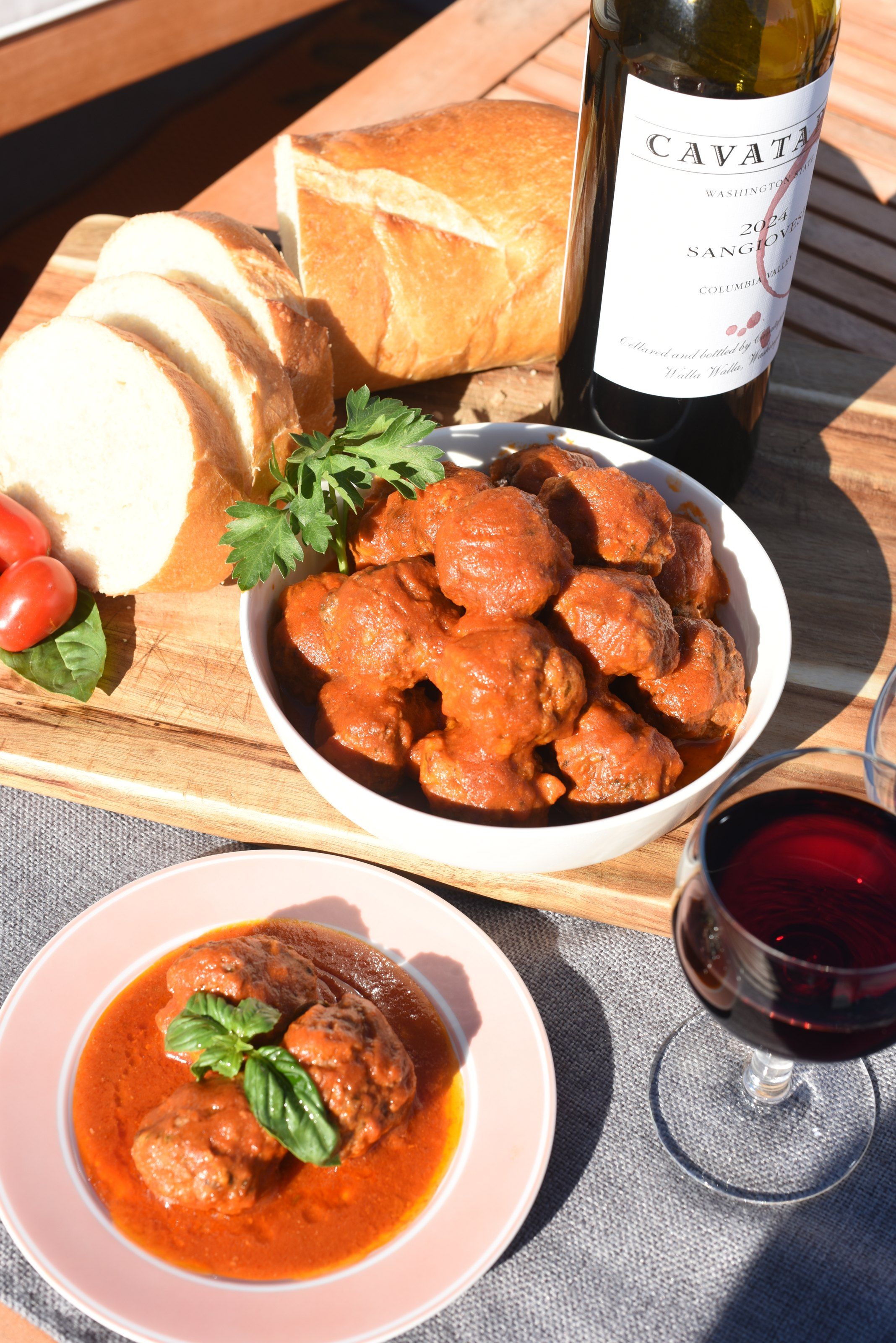 A bowl of meatballs in tomato sauce with basil, a plate of two meatballs, bread, cherry tomatoes and red wine on an outdoor table
