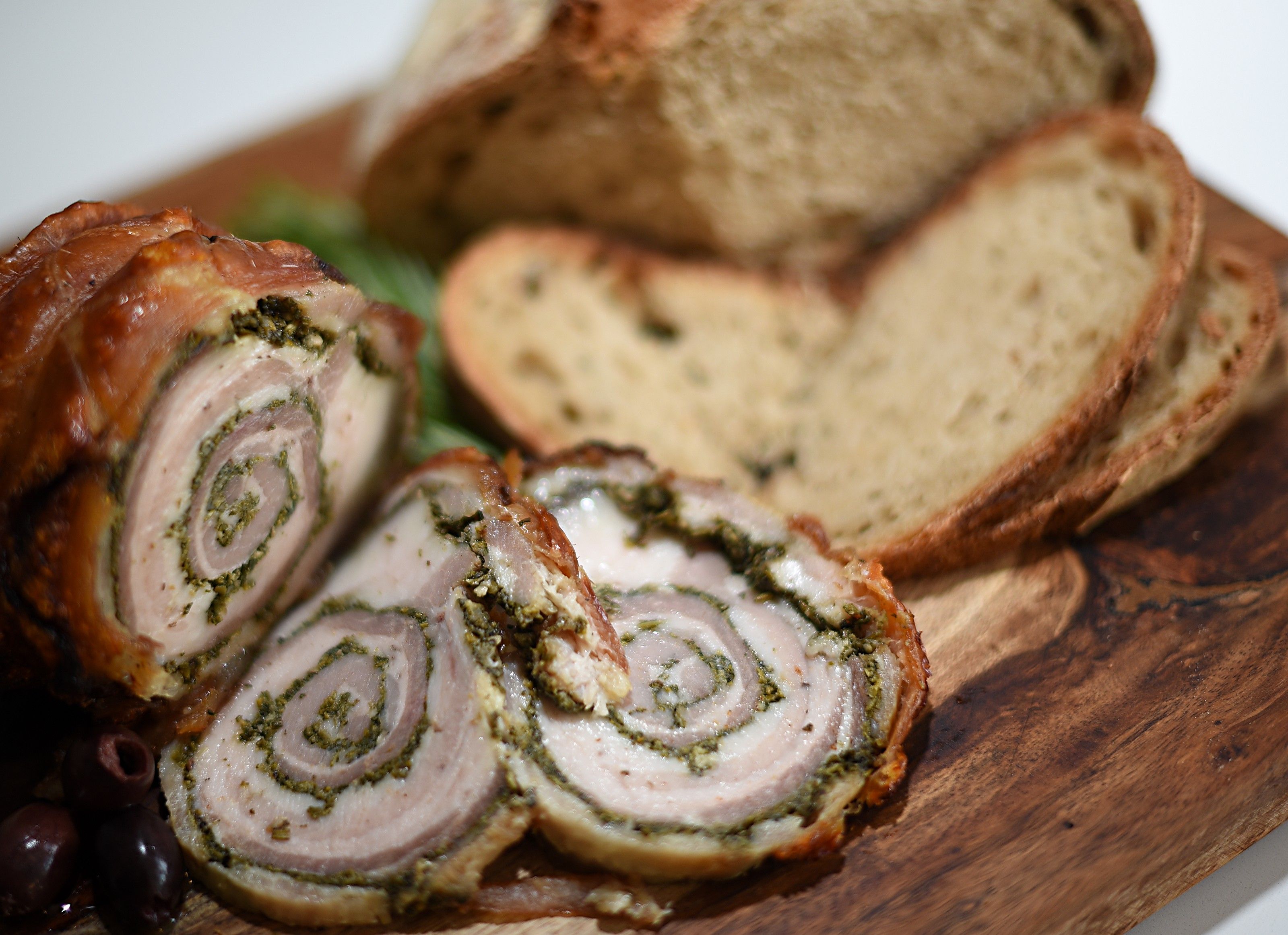 Slices of porchetta rolled around a green herb filling, with sliced bread and black olives on a wooden board
