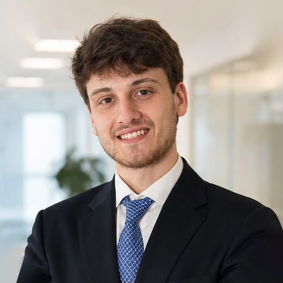 Alex in a dark suit and blue tie, smiling, in a bright office