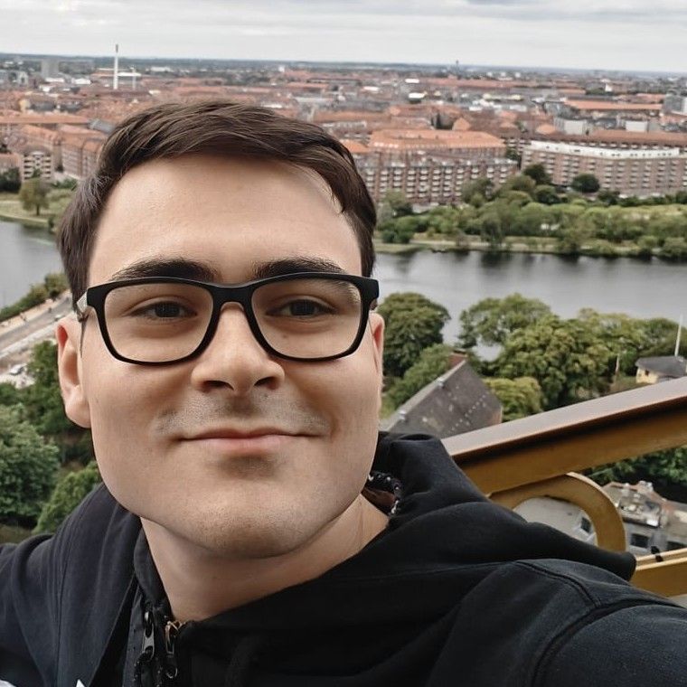 Michele, smiling, in glasses and a black hoodie, with a city and a lake behind him