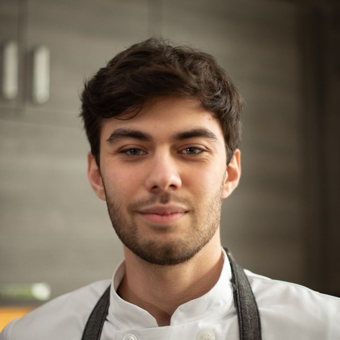 Chef Nicholas Olson in a white chef's jacket and grey apron, smiling, in a kitchen
